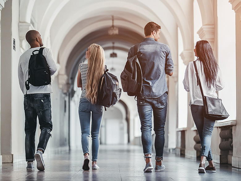 Four young adult students, each carrying a backpack, walking down a university hallway together with their backs toward the camera.